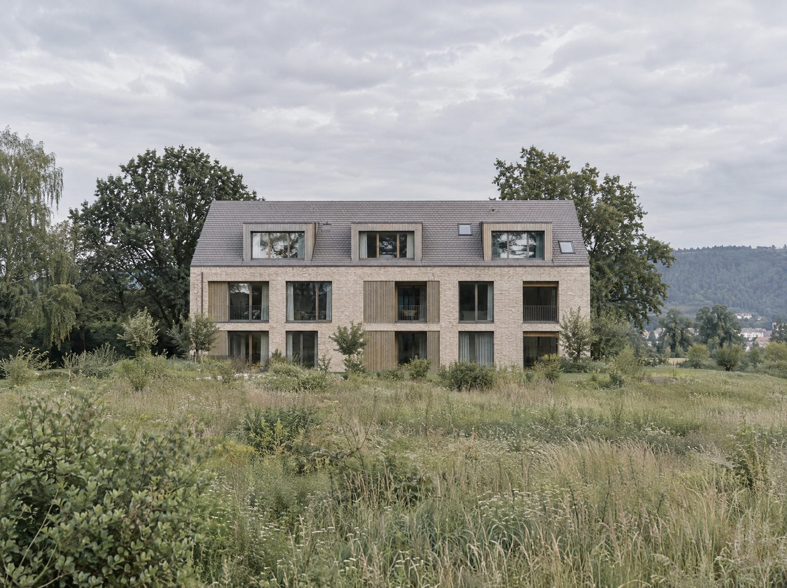 Mehrfamilienhaus aus Klinker mit Holzelementen und Gauben, in einer Wiese vor bewaldeten Hügeln